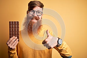 Irish redhead man with beard eating a sweet chocolate bar over yellow isolated background happy with big smile doing ok sign,