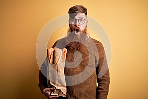 Irish redhead man with beard holding groceries paper bag of bread over yellow background scared in shock with a surprise face,