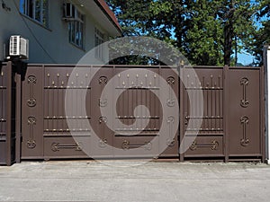 Iron brown automatic gates and a wicket with a forged gilded pattern on them are illuminated by the sun