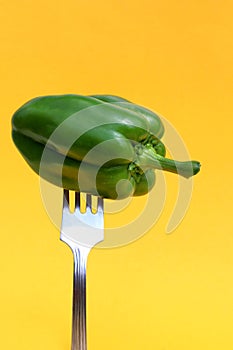 An iron fork is stuck in a green bell pepper, on a yellow background.