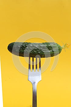 An iron fork is stuck in a green cucumber on a yellow background.