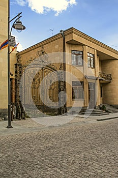 Iron gate with monogram next to a building made of light tuff with a wrought iron balcony in a niche on Abovyan street in Gyumri