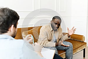 An irritated African American emotionally talks about problems to a psychologist at a session, a confused unhappy young