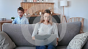 Irritated busy freelancer female working laptop sitting couch surrounded by noise running children