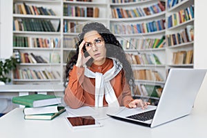 Irritated public library worker rolling eyes and leaning head on hand by desk with computer and books. Annoyed woman