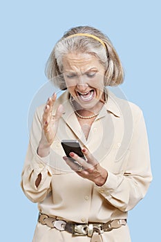 Irritated senior woman reading text message on cell phone against blue background