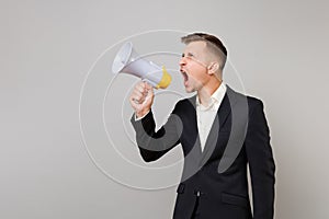 Irritated young business man in classic black suit, shirt looking aside, screaming on megaphone isolated on grey