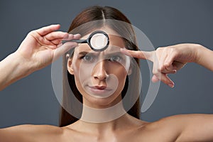 Irritated young lady examining skin with magnifying glass and frowning