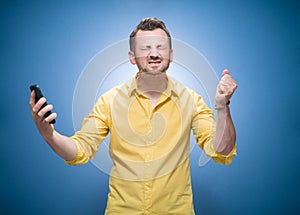 Irritated young man talks on the phone over blue background, dresses in yellow shirt. Annoyed guy talking. Stressed person using