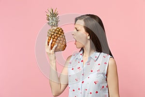 Irritated young woman in summer clothes holding in hand fresh ripe pineapple fruit isolated on pink pastel wall