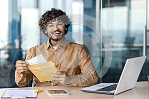 Islamic guy sitting by worktable with portable gadgets and opening envelope with paperwork correspondence. Happy