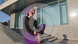 Islamic muslim woman student girl in hijab sitting on background of city building outdoors chatting on mobile phone