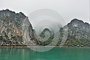 Islands in Halong Bay in the fog. Calm emerald water and a narrow strip of sand on the shore.