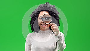 Isolated black female on a green screen smiling and holding a magnifier in front of her left eye