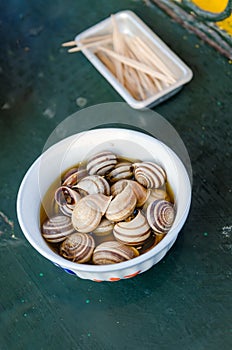 Isolated boiled or cooked snails with striped shells in smaall bowl with toothpicks on table, Marrakesh, Morocco