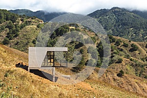An isolated modern architectural structure stands in contrast to the vast, hilly terrain of a mountainous landscape. At San