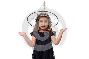 Isolated portrait of pretty little witch girl wearing wizard hat, dressed in stylish carnival dress, gesturing, holding a copy