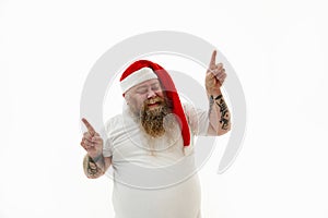 Isolated portrait of an overweigh smiling bearded man in Santa hat with tattooed arms on the head pointing his fingers up