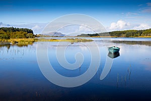 An isolated rowing boat sits on a calm blue coloured lake with distant mountains and forestry.