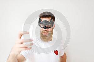 Isolated shot of attractive male with thick long beard and mustache covers face with batman mask, looks at smart phone