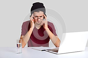 Isolated shot of curly male worker tries to concetrate on work, sits at desktop in front of opened laptop, keeps fingers on