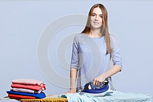 Isolated shot of hard working female busy with house work, stands in laundry, irons clothes with electric iron, isolated over blue