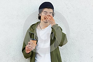 Isolated shot of sleepy curly man keeps hand on nose, dressed in stylish hat and anorak, holds takeout coffee, wants to have rest