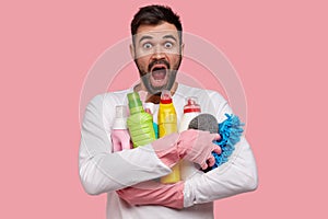 Isolated shot of stupefied bearded emotional man carries bottles of detergents closely, stares with unbeliveable
