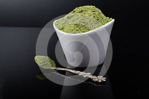 An isolated tablespoon of dried organic wheat grass and spirulina powder, on white rustic background