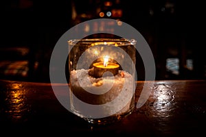 Isolated tealight inside a glass illuminating a dark scene and reflections on a wooden table and a nice bokeh background