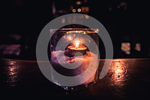 Isolated tealight inside a glass illuminating a dark scene and reflections on a wooden table and a nice bokeh background