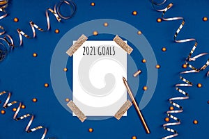 Isolated white card with 2021 goals wording and golden pen with festive golden ribbons and confetti. Flat lay background
