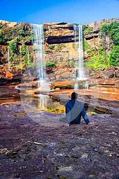 Isolated young man sitting near waterfall falling from mountain top with reflection and blue sky
