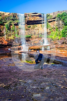 Isolated young man sitting near waterfall falling from mountain top with reflection and blue sky