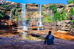 Isolated young man sitting near waterfall falling from mountain top with reflection and blue sky