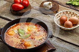 Israeli fried eggs shakshuka with cheese in a frying pan near products on a wooden table.