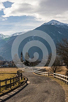 Italian Alps, asphalt road fenced with a wooden fence leading to alpine village