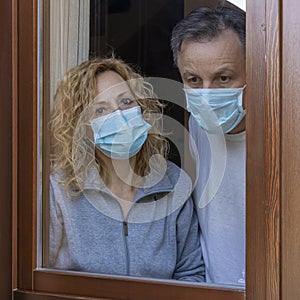 Italian couple with protective mask at the window looks out with a dejected expression because of the quarantine from covid-19