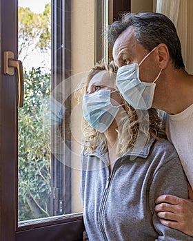 Italian couple with protective mask at the window looks out with a dejected expression because of the quarantine from covid-19