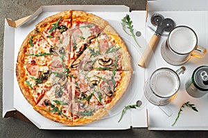 Italian fast food. Delicious hot pizza in a box with ham, champignons and glass of beer sliced and served on brown table, close up