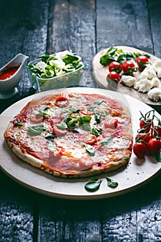 Italian food, cuisine. Margherita pizza on a black, wooden table with igredients like tomatoes, salad, cheese, mozzarella, basil.