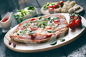 Italian food, cuisine. Margherita pizza on a black, wooden table with igredients like tomatoes, salad, cheese, mozzarella, basil.