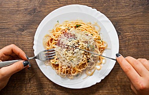 Woman hand holding fork and knife with spaghetti bolognese in white plate on wooden table. top view