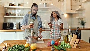 Italian man, chef cook using hand blender, preparing a meal while his girlfriend, young woman in apron pouring two