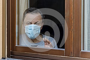 Italian man with protective mask, forced to stay at home due to coronavirus covid-19, disconsolate looks out the window