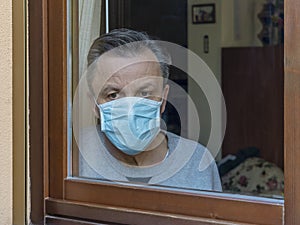 Italian man with protective mask, forced to stay at home due to coronavirus covid-19, disconsolate looks out the window
