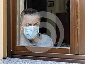 Italian man with protective mask, forced to stay at home due to coronavirus covid-19, disconsolate looks out the window