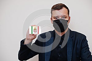 Italian man wear black formal and protect face mask, hold Apulia flag card isolated on white background. Italy regions coronavirus