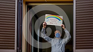 An italian middle-aged couple looks out the window with protective masks and exposes the sign `Andra` tutto bene`