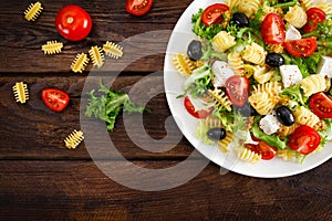 Italian pasta salad with fresh tomato, cheese, lettuce and olives on wooden background. Mediterranean cuisine. Cooking lunch. Hea